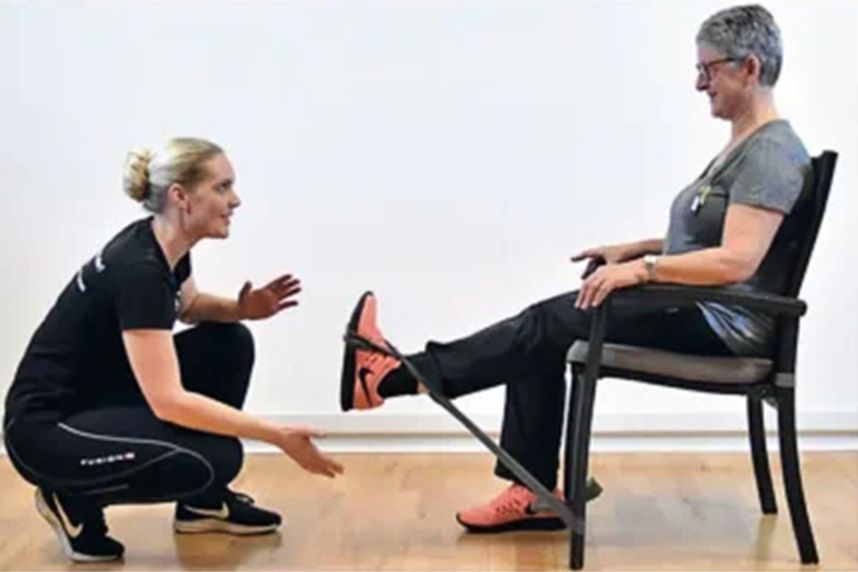 Physiotherapeutin leitet eine ältere Frau beim Beinstreck-Übung im Sitzen auf einem Stuhl an